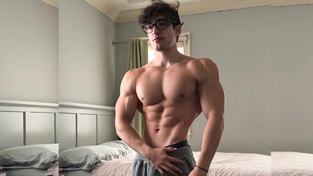 Chris Aronis with glasses posing in a bedroom, one hand at his waistband beside a white bed and blue wall.