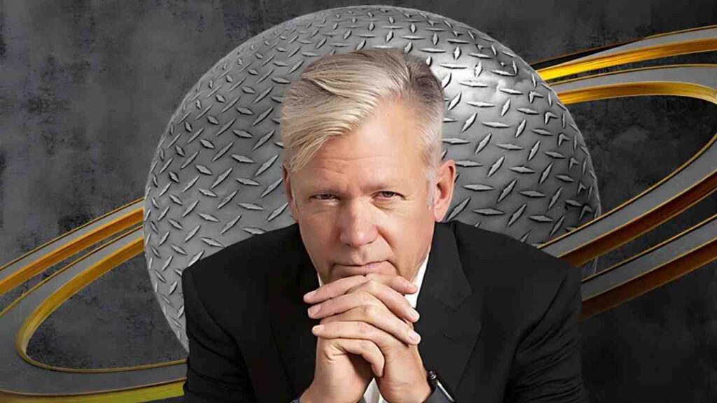 Chris Hansen in a black suit with blond hair rests his clasped hands under his chin before a metallic diamond-plate backdrop.