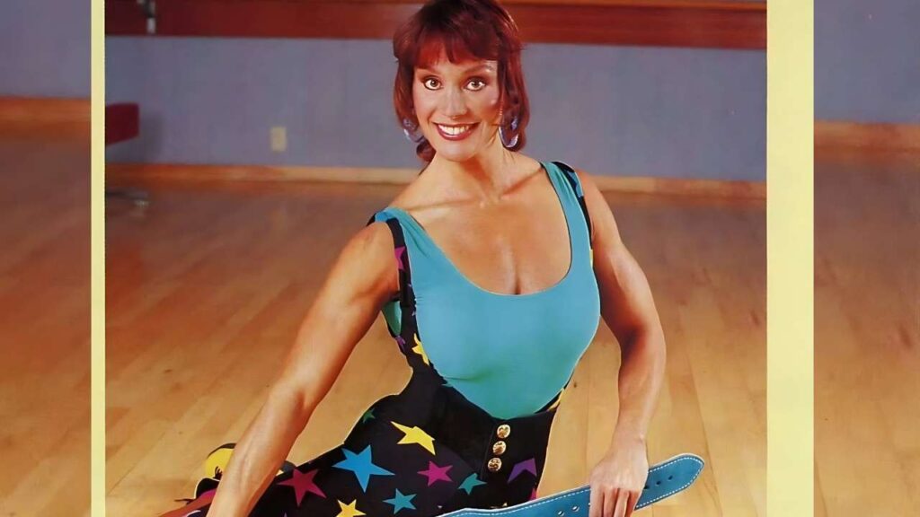 Cory Everson with short red hair in turquoise top and starry skirt sits on a wooden dance floor, holding a blue skateboard.
