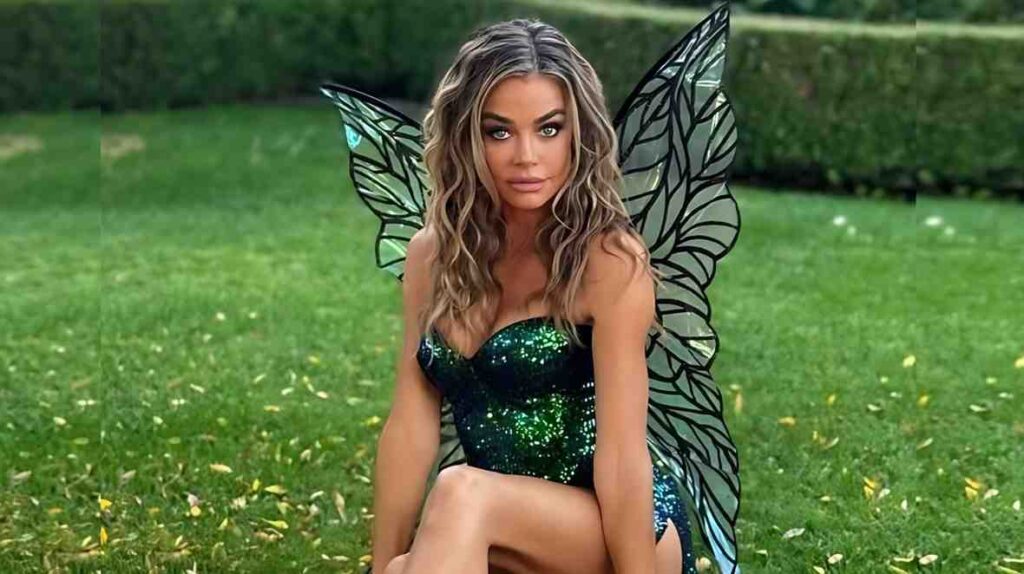Denise Richards in a sparkling emerald strapless dress with black-veined fairy wings sits on grass in a lush garden.