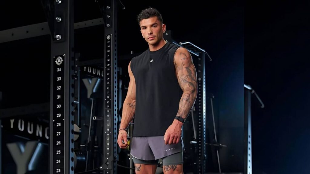 Dorian Vranjes in a black sleeveless shirt and gray shorts poses in a gym beside a squat rack, tattoos on his arms.