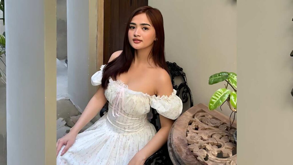 Eleiza Jordanne Abejon Serante in an off-shoulder white floral dress sits on an ornate black chair beside a table with a green potted plant.
