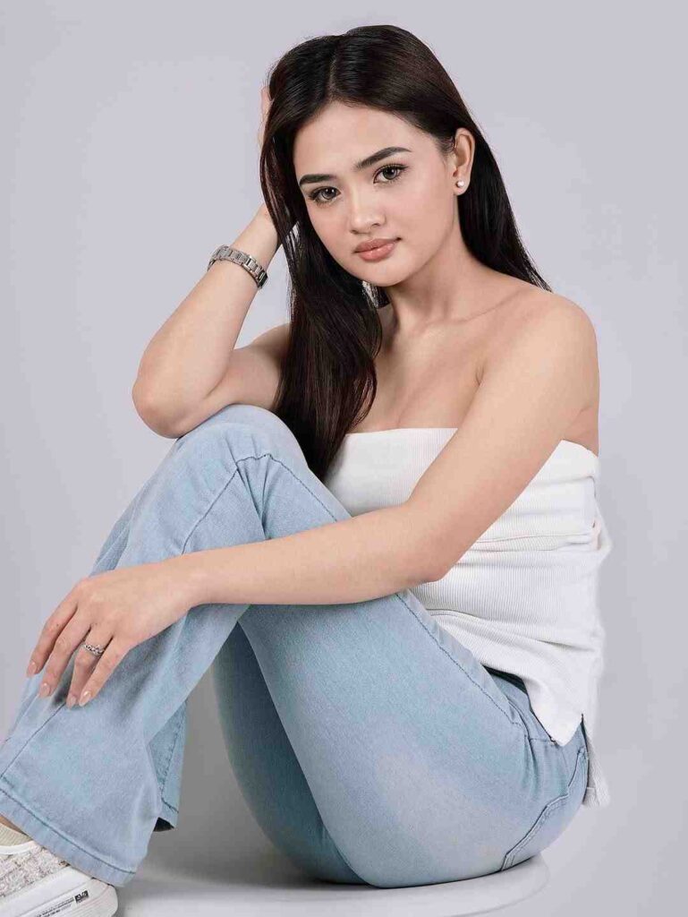 Eleiza Jordanne Abejon Serante sits against a gray backdrop in a white off-shoulder top and light jeans, looking at the camera.
