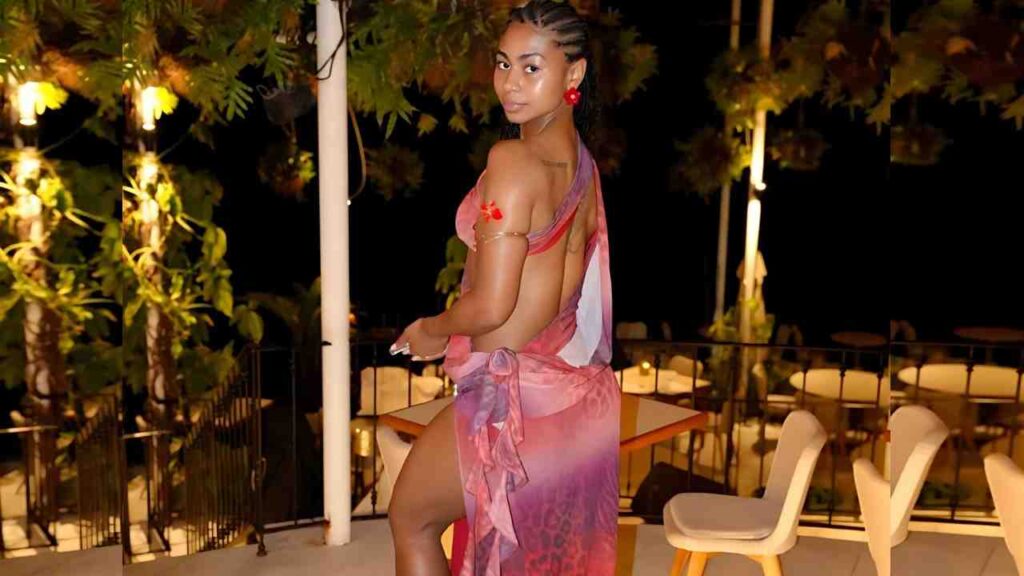 Elizabeth Mira in a pink-purple gradient wrap dress with braided hair stands on a night-lit outdoor balcony, looking over her shoulder.
