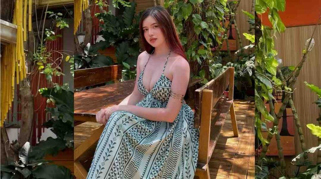 Elyza Macapagal in a blue geometric dress sits on a wooden bench on a sunny tropical patio, surrounded by lush plants.