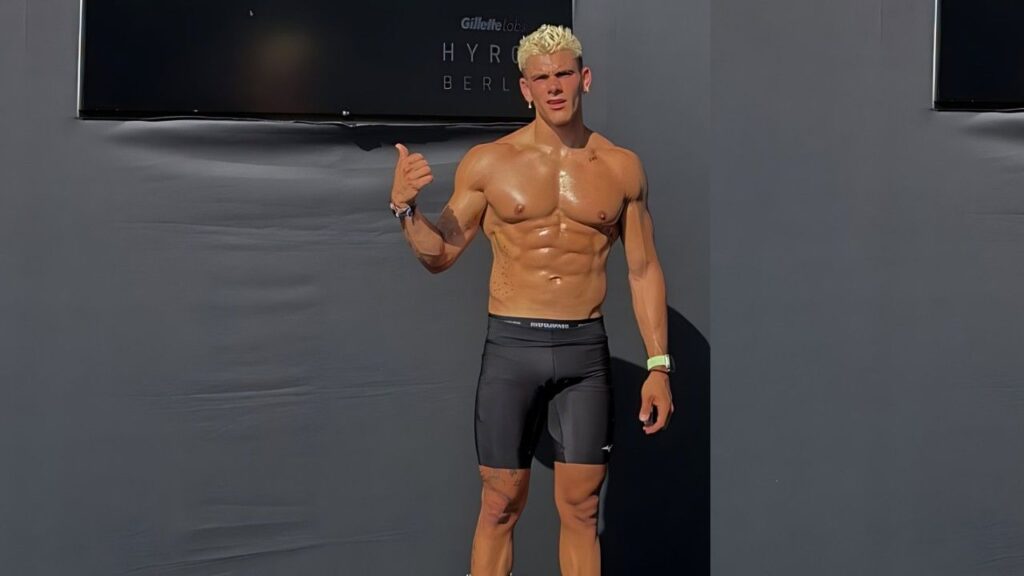 Filippo Tenca with bleached blond hair stands against a gray wall in black compression shorts, giving a thumbs-up.
