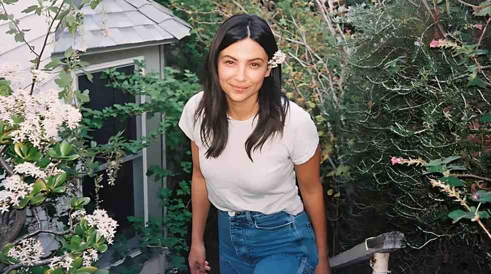 Floriana Lima with dark hair in a white tee and jeans stands in a lush garden, a white flower behind her ear, smiling softly.