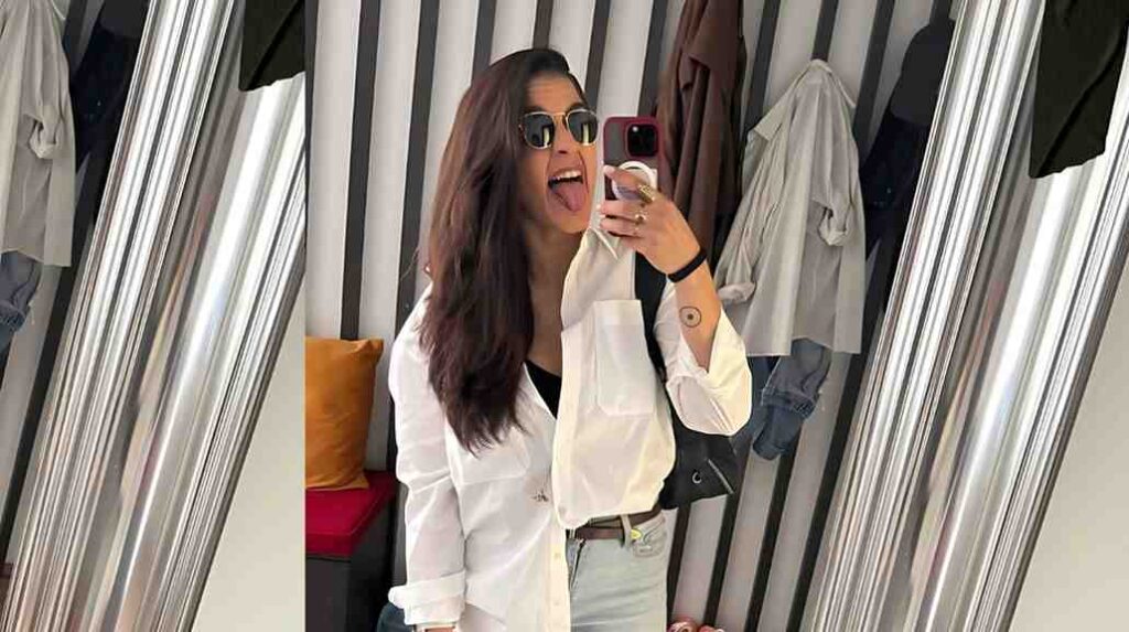Franca Molluzzo in a dressing room snaps mirror selfie, tongue out, white shirt over black top with jeans, holding pink phone.