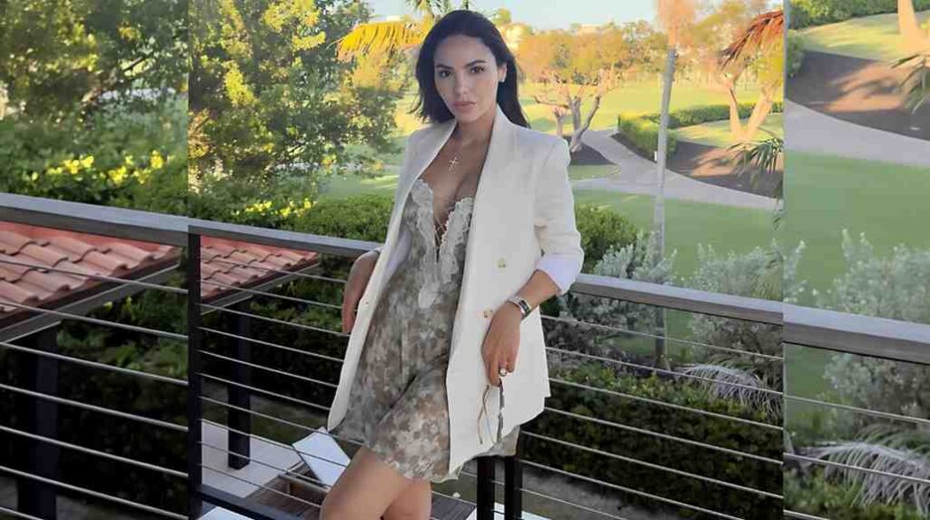 Francesca Frigo stands on a balcony in a white blazer over a gray mini dress, holding sunglasses against sunny garden backdrop.
