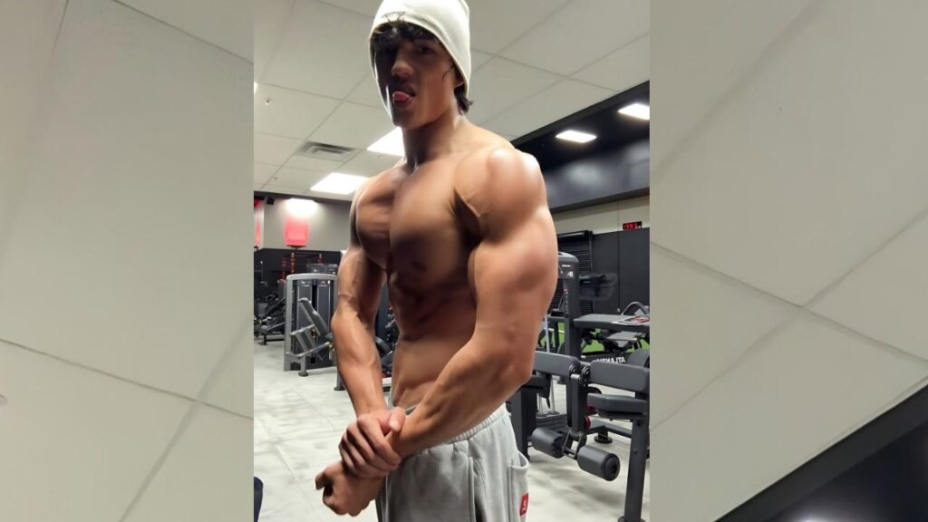 Gabe Deutsch in a light beanie sticks out his tongue while flexing his biceps in a gym, wearing gray sweatpants.