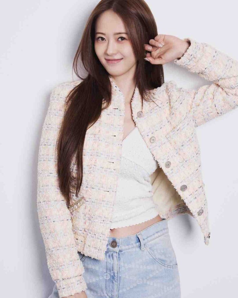 Go Ara with long chestnut hair wears a pastel tweed jacket over a white top and light jeans, posing with one hand.