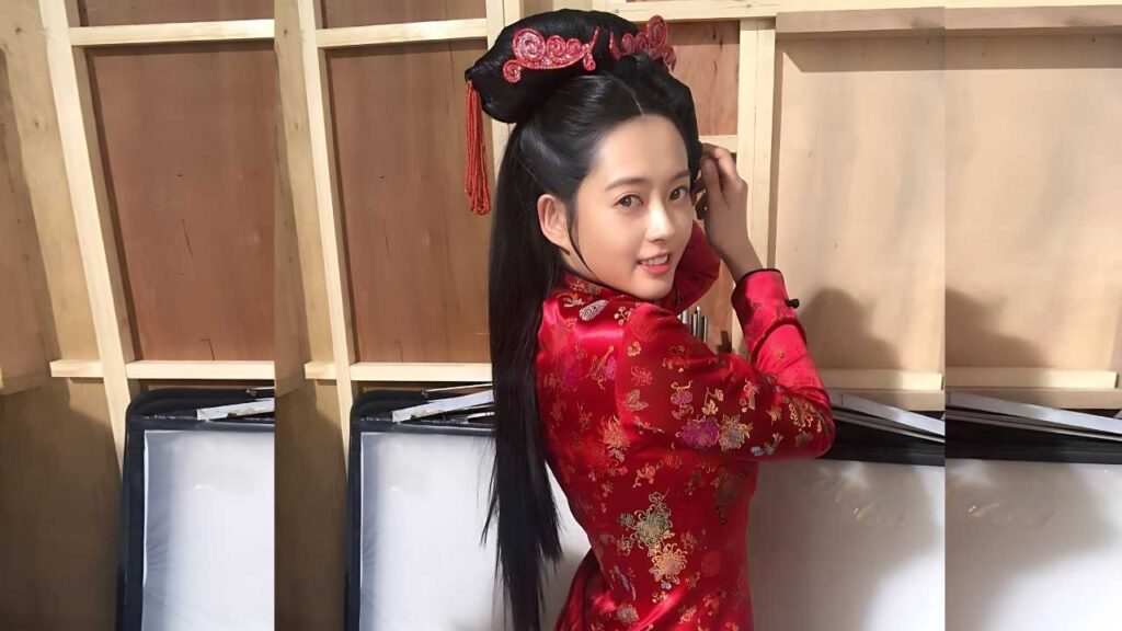 Go Ara in a red embroidered cheongsam with an ornate headdress, smiling as she adjusts her hair in a wooden studio.