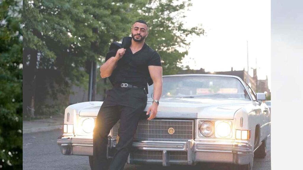 Haddy Abdel in a black polo and black pants leans on a white vintage Cadillac, jacket slung over his shoulder, along a sunlit street with trees.