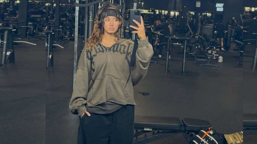 Ingrid Munn in an oversized gray hoodie and headband takes a mirror selfie in a gym, with benches and weight racks visible.