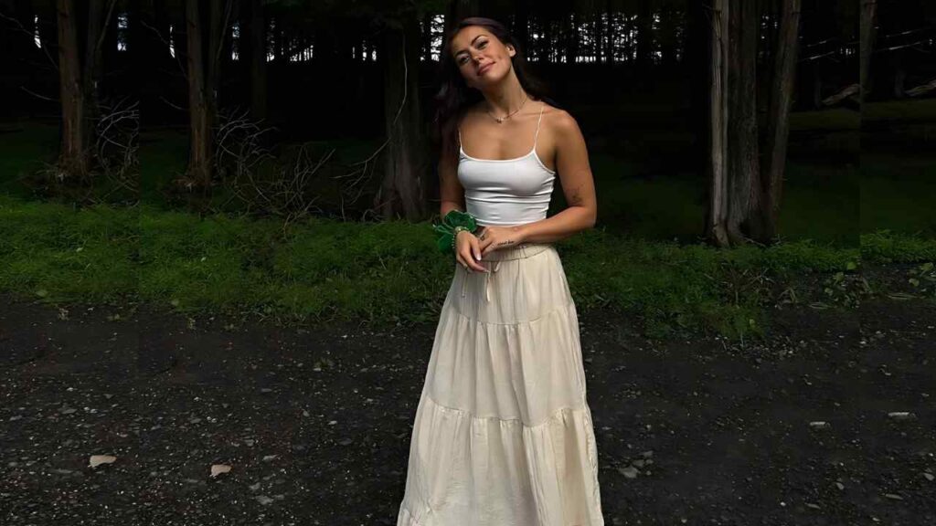 Iris Dailey in white tank and cream maxi skirt stands on a gravel path in a forest, hands clasped, green scrunchie on wrist.