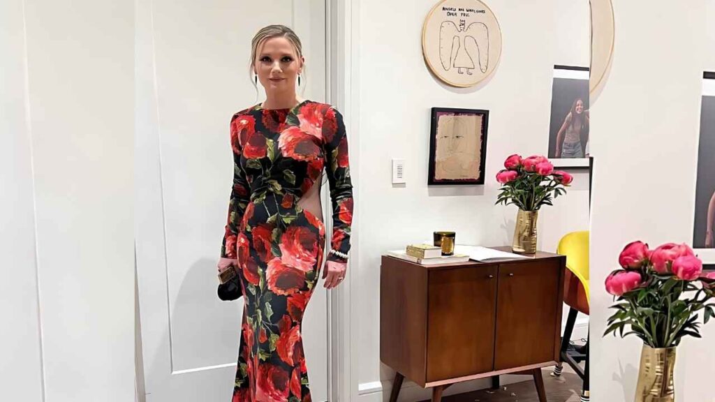 Jennifer Nettles in a bold red floral gown poses in a bright gallery with a wood sideboard, pink peonies, and framed art.