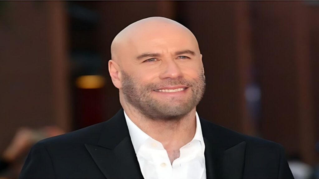 John Travolta with a trimmed beard in a black suit and white shirt, indoors with a blurred warm-toned background.