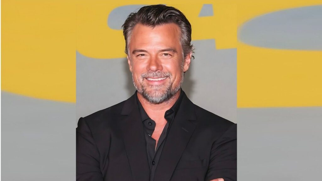 Josh Duhamel with salt-and-pepper hair and beard in a black suit and open-collar shirt, posed against a bright yellow-gray backdrop.