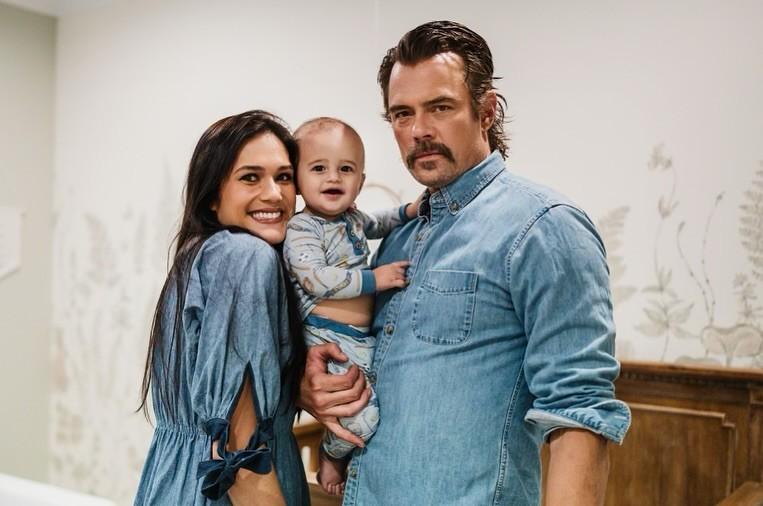 Smiling trio indoors: Josh Duhamel wife with long dark hair in a blue denim dress, Josh Duhamel in a denim shirt holding their baby
