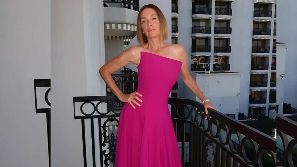 Julianne Nicholson in a magenta one-shoulder gown poses on a balcony, hand on hip, with a modern hotel backdrop.
