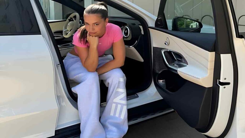 Justine Antoine in pink top and lavender sweatpants sits in the open door of a white luxury car, chin on hand, gazing at camera.