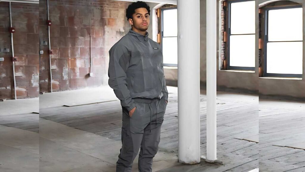 Justmerk in a gray tracksuit stands with hands in pockets inside a brick-walled warehouse with windows and white columns.