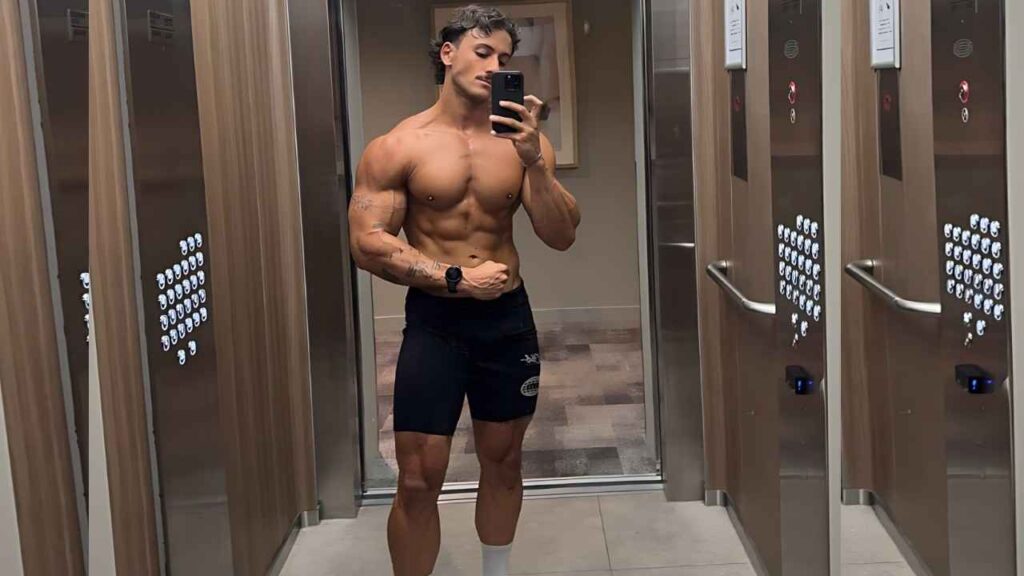 Kalani James Lenehan in a wood-paneled elevator flexes his bicep while taking a mirror selfie, wearing black shorts.