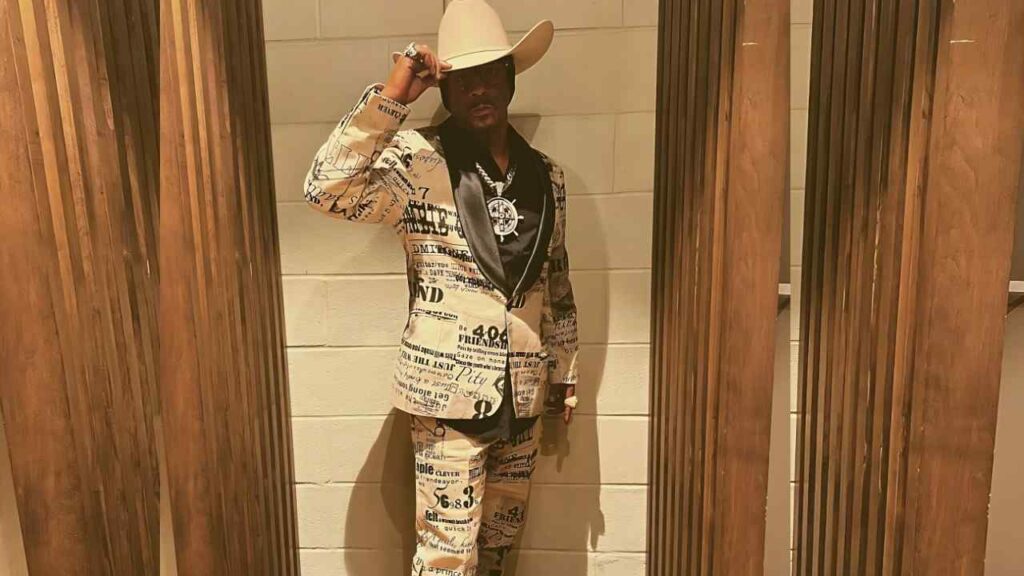 Katt Williams in beige newspaper-print suit and black shirt, wearing a cowboy hat and necklace, tipping his hat between wooden frames.