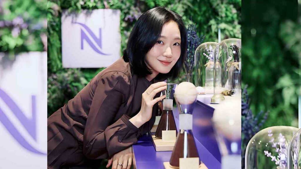 Kim Go-eun with short black hair and a brown top leans over a purple display, inspecting grooming tools on wooden stands.
