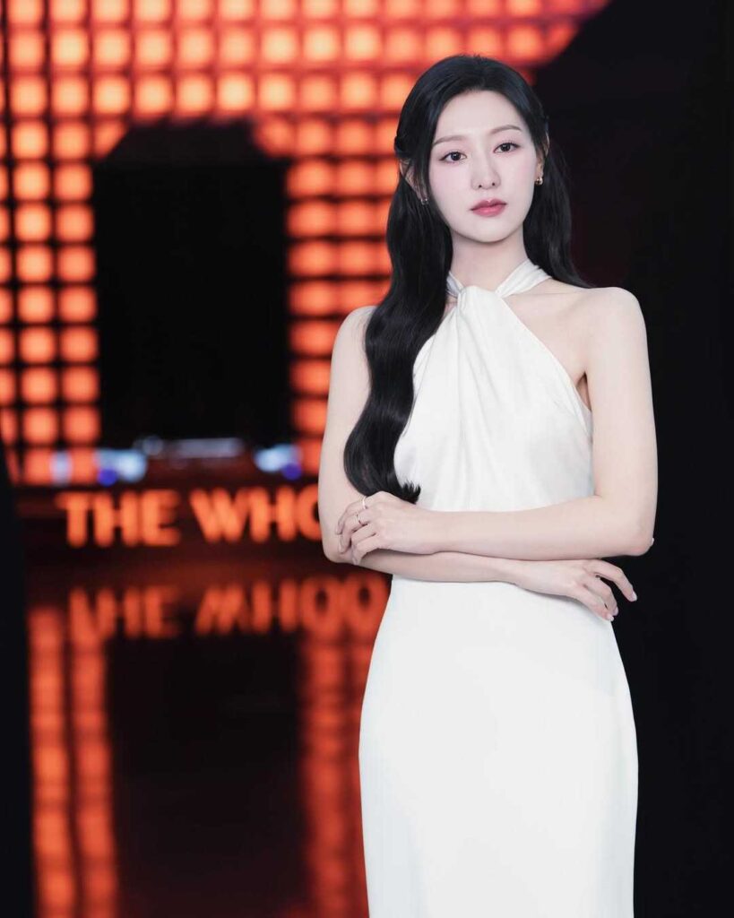 Kim Ji-won height with long black hair in a white halter-neck gown stands with arms crossed before a red, grid-lit backdrop.