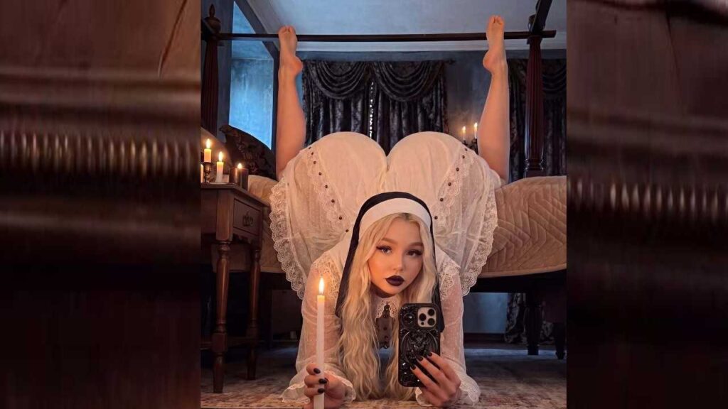 Kristina Musatova in white lace nun costume lies on the floor, candle lit, holding a phone, legs raised over a four-poster bed.