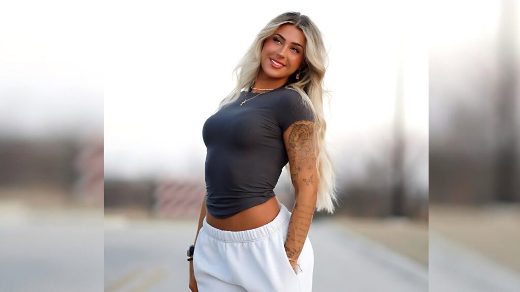Lauren Wagner in a tight gray crop top and white joggers stands on a blurred outdoor road, showing left-arm tattoos.