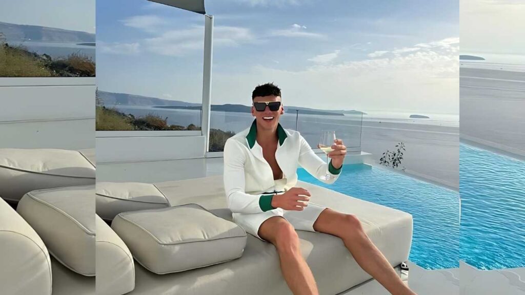 Lewis McGarry in a white blazer with green trim lounges by a pool, sunglasses on, holding a glass of white wine, sea behind.