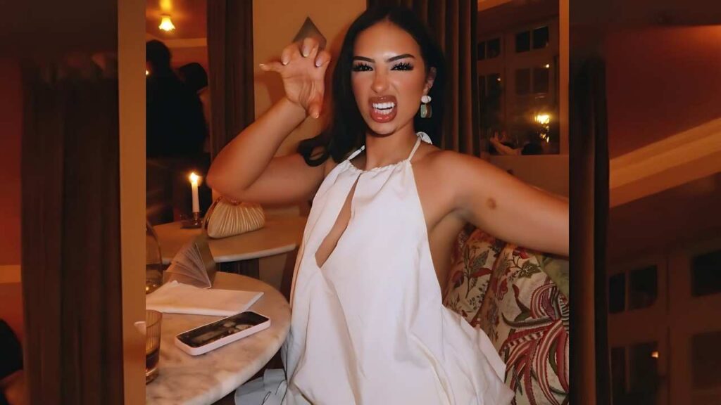 Line Souaidi in a white halter dress scrunches her face and claws toward the camera, seated at a candlelit marble table with a phone nearby.