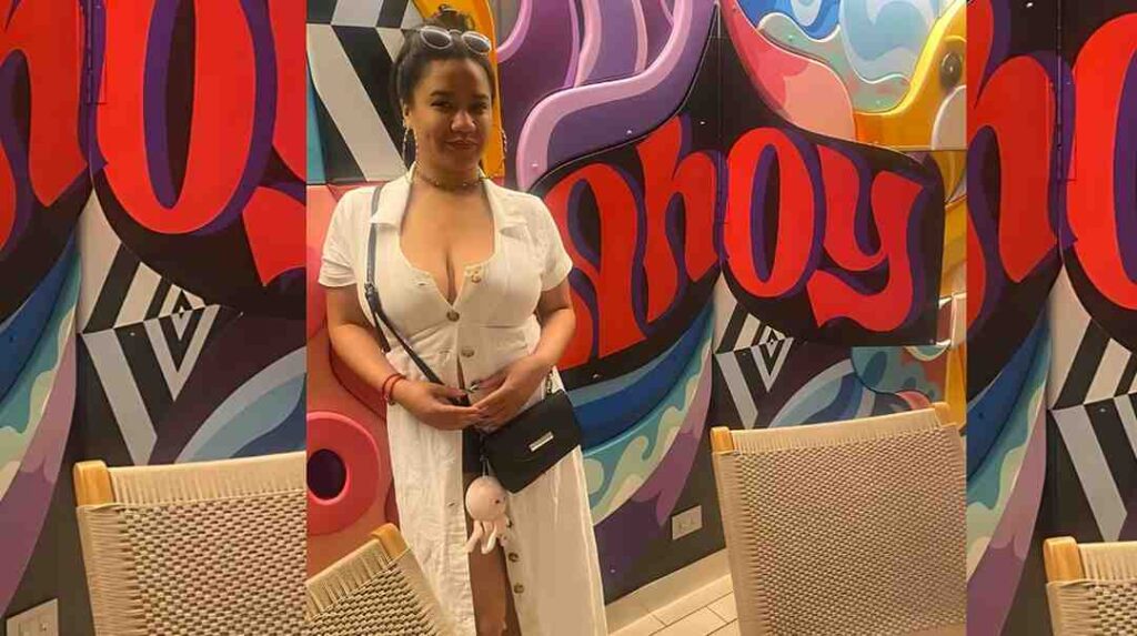Loni Legend in a white dress stands before a colorful graffiti wall, with a black crossbody bag and plush keychain; wicker chairs in foreground.