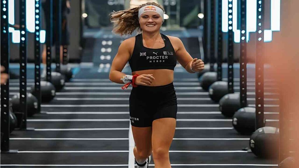 Lucy Procter runs through a rack-lined gym, wearing black sports bra and shorts, white headband and red wristband.