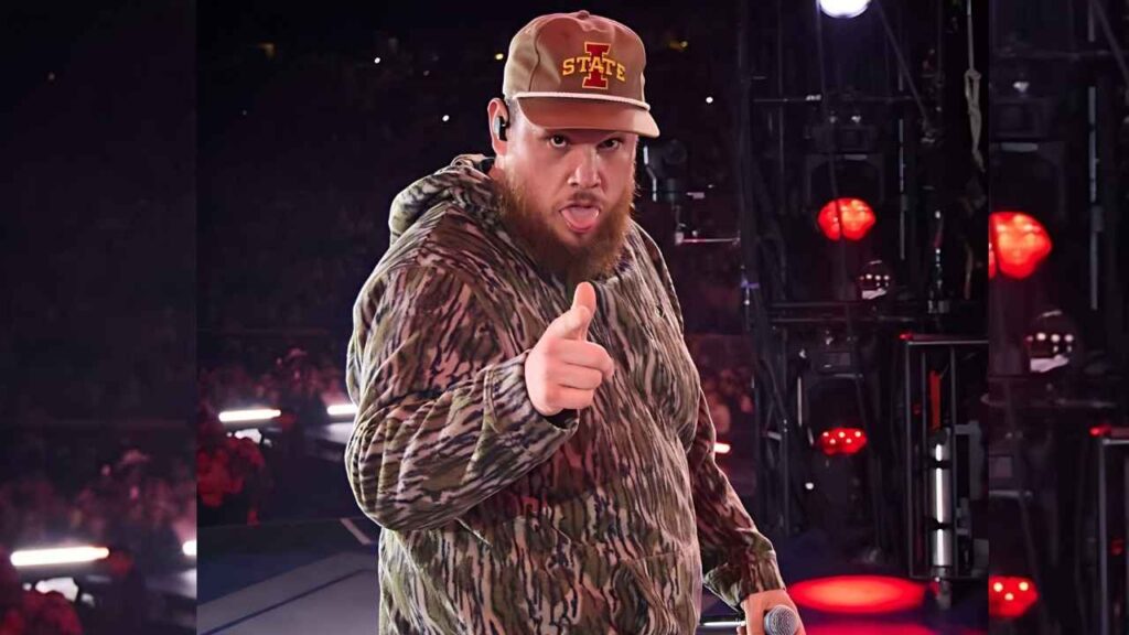 Luke Combs in camo hoodie and brown cap points at the camera on a stage, holding a mic, red lights and cheering crowd behind.