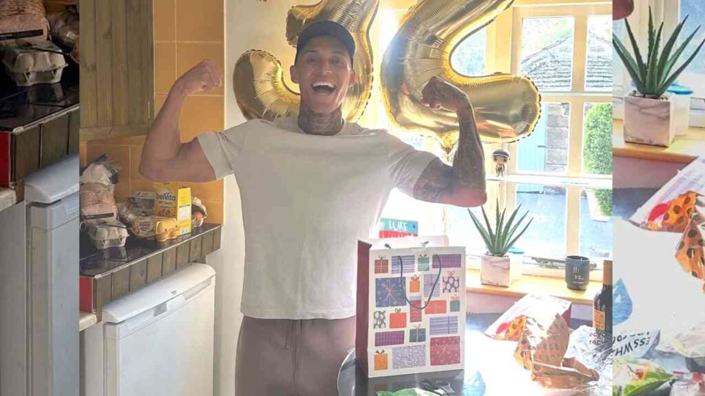 Luke Stoney in white tee and cap flexes tattooed arms in a kitchen, gold number balloons behind him, gift bag on table by window.