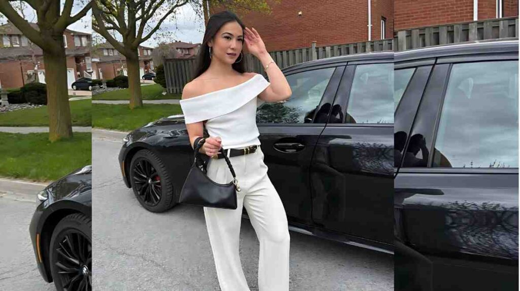 MDJ Fitness in an off-shoulder white outfit with a black belt poses beside a sleek black SUV, carrying a black handbag.