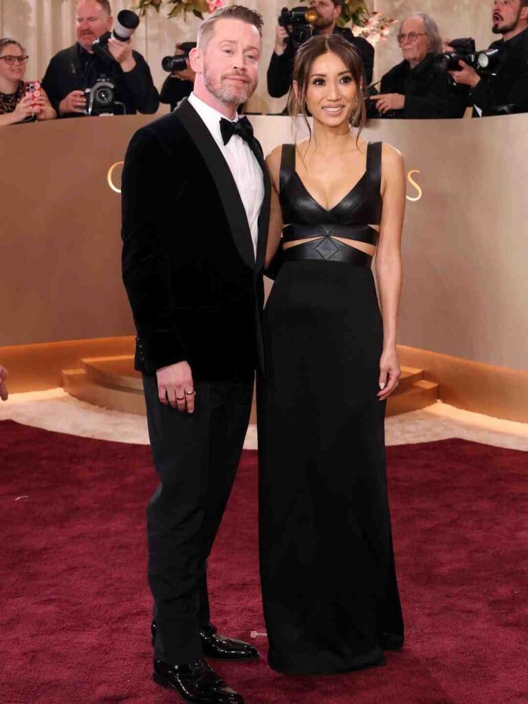 Macaulay Culkin on red carpet in velvet tuxedo with bow tie and Macaulay Culkin wife in a black cutout gown, photographers behind them.