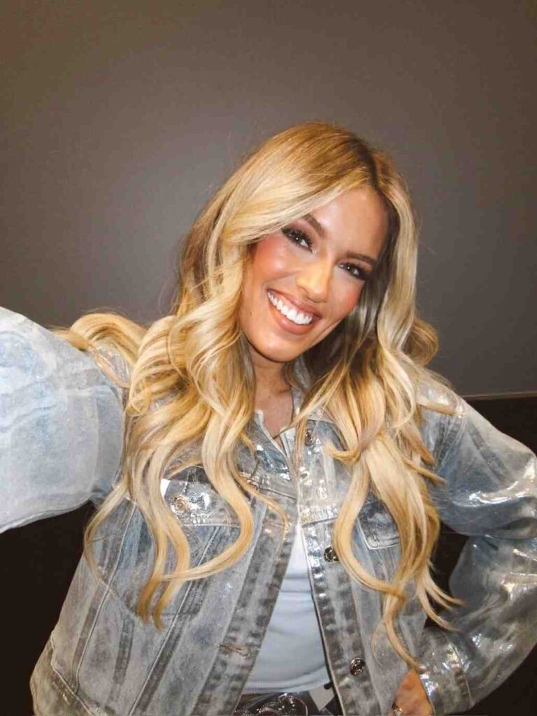 Mackenzie Carpenter with long, wavy blonde hair wearing a light-wash denim jacket, posing for a selfie against a gray wall.