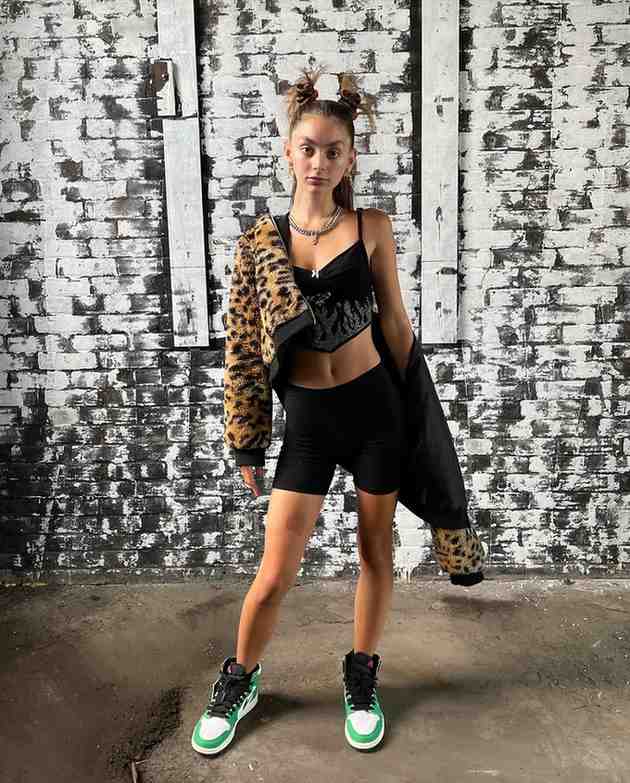 Mackenzie Shirilla today stands before a weathered white brick wall, wearing a leopard-print jacket draped over a black crop top and shorts, bright green-and-white sneakers, and space buns.