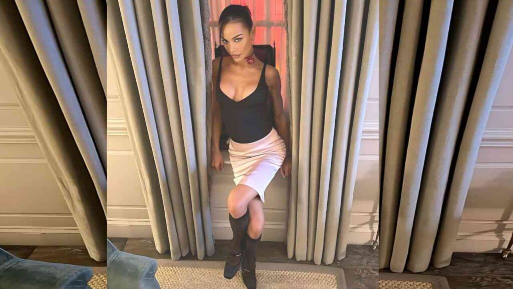Madison Sarah stands between beige curtains, wearing a black sleeveless top, pale pink pencil skirt, and knee-high boots.