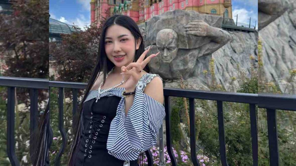 Manila Bianco with long black hair stands by a railing, making a peace sign, with a rock sculpture and castle behind.