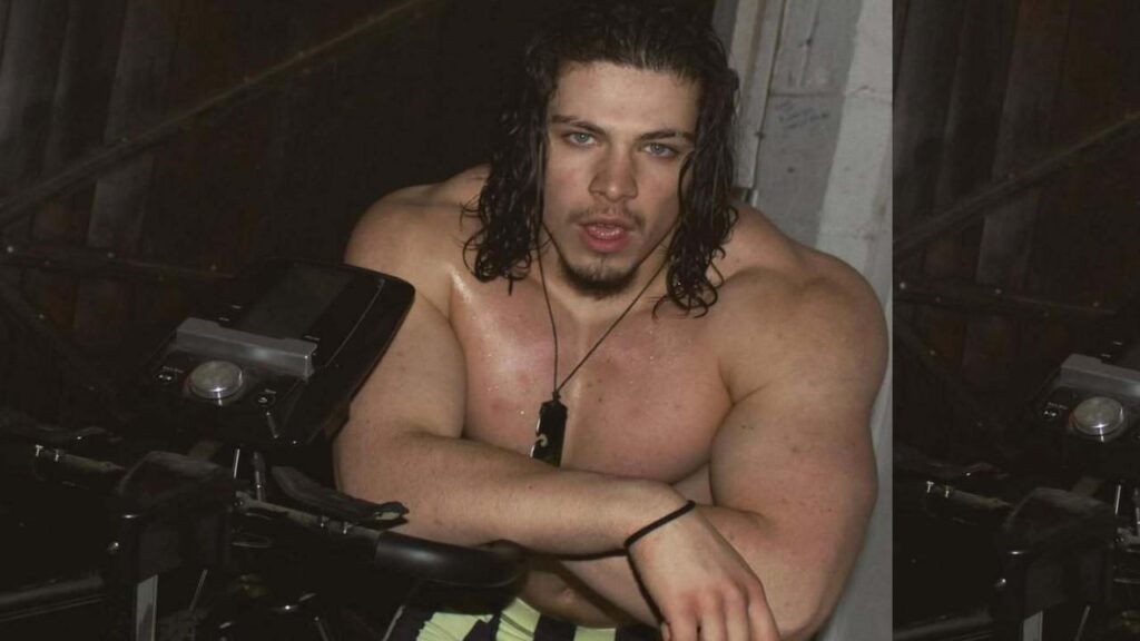 Marcus Jason Brown with long wet hair and a dog tag necklace rests on a motorcycle in a dim wood-walled garage.