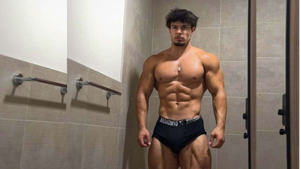 Mike Aguilera in black briefs poses in a tiled locker room, wearing a dog-tag necklace with a serious expression.
