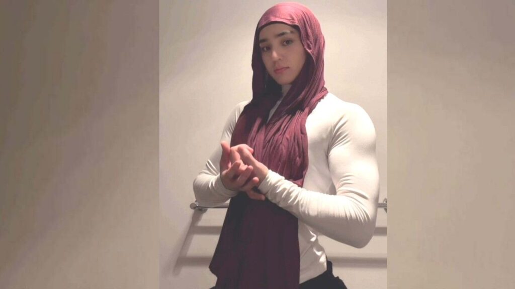 Nasrin Yawari in a burgundy hijab and white long-sleeve top stands against a beige wall, hands clasped, calm expression.