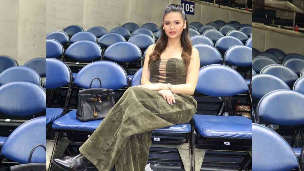 Nathalie Colipano with long wavy hair sits among blue stadium seats, wearing an olive-green strapless dress and a black handbag.