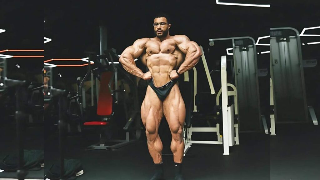 Niall Darwen bodybuilder in a dim gym wearing black posing trunks and glasses, strikes a dramatic double-biceps pose amid gym equipment.