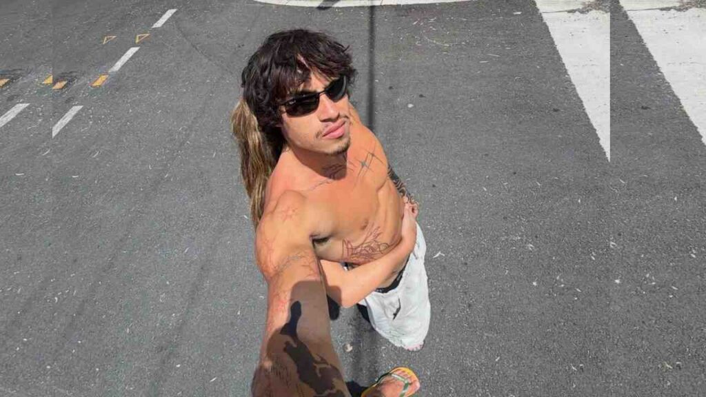 Noah Rolette with long mullet and sunglasses, tattooed arms, poses on a street near a crosswalk; shadow in foreground.